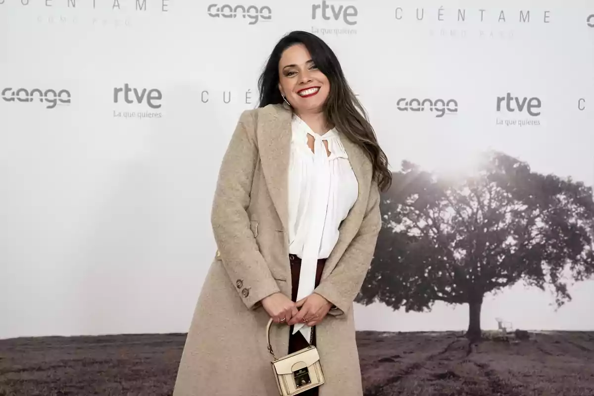 Mujer sonriendo con abrigo beige y bolso pequeño posando frente a un fondo promocional de la serie Cuéntame cómo pasó Mujer sonriendo con abrigo beige y bolso pequeño posando frente a un fondo promocional de la serie Cuéntame cómo pasó