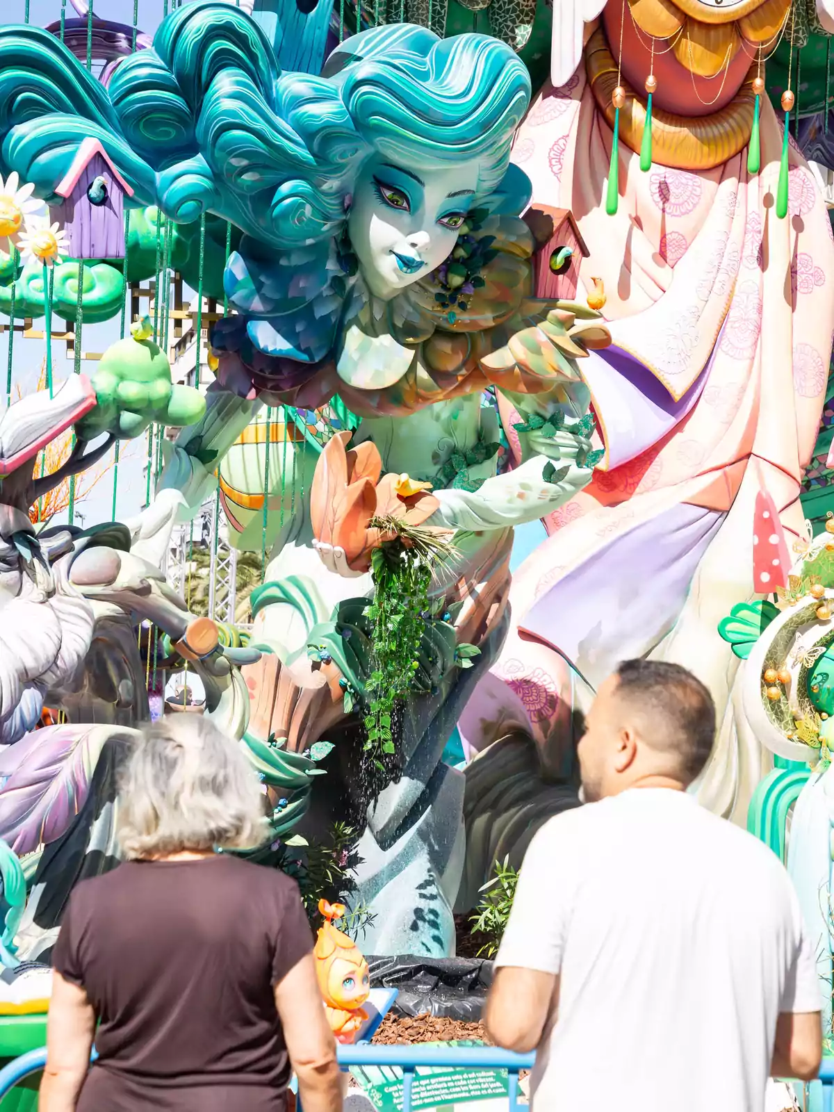 Dos personas observan una colorida escultura de una figura femenina fantástica con cabello azul y detalles florales en un evento al aire libre. Dos personas observan una colorida escultura de una figura femenina fantástica con cabello azul y detalles florales en un evento al aire libre.