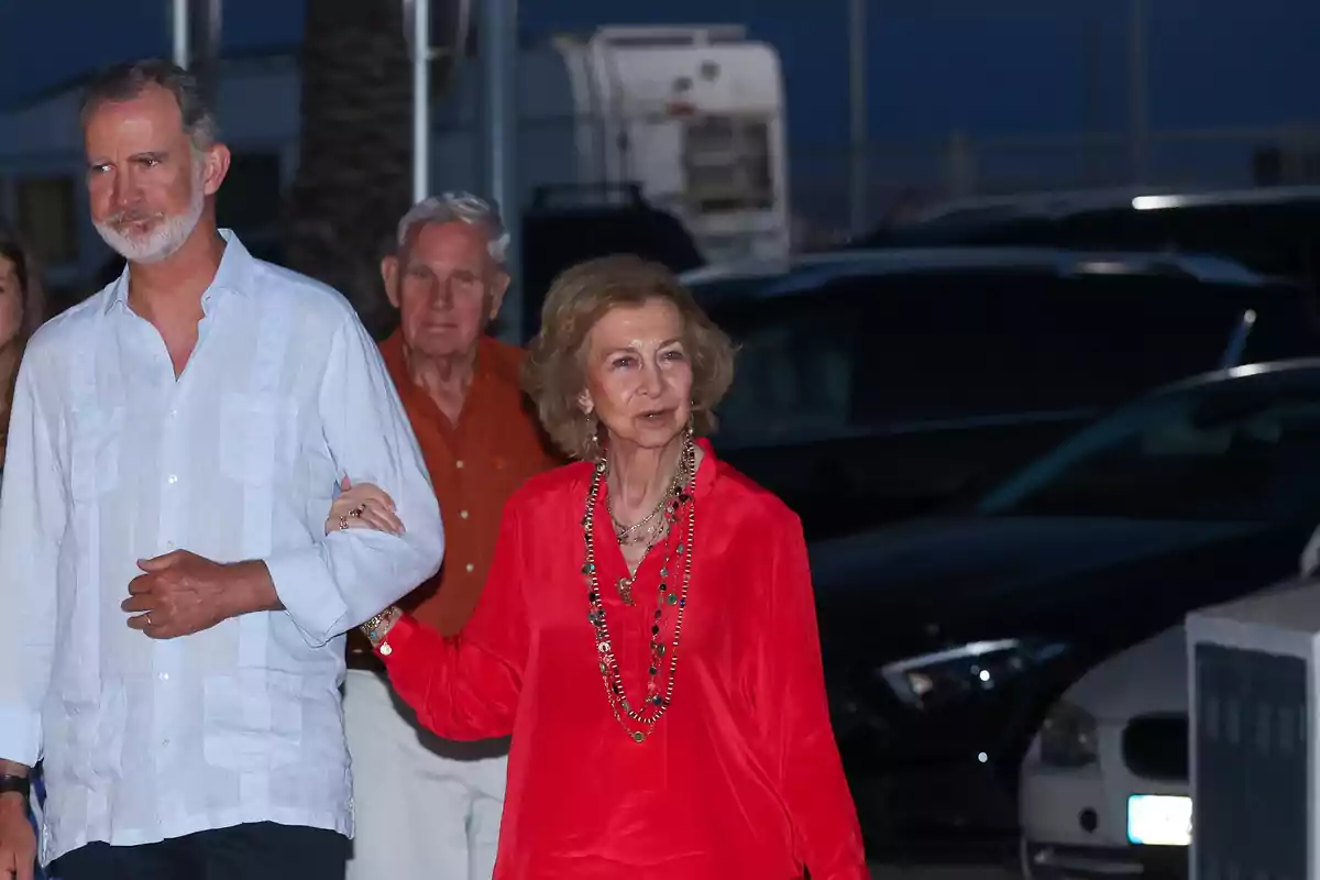 Una mujer mayor vestida con blusa roja y collar largo camina tomada del brazo de un hombre de barba y camisa clara en un estacionamiento al anochecer Una mujer mayor vestida con blusa roja y collar largo camina tomada del brazo de un hombre de barba y camisa clara en un estacionamiento al anochecer