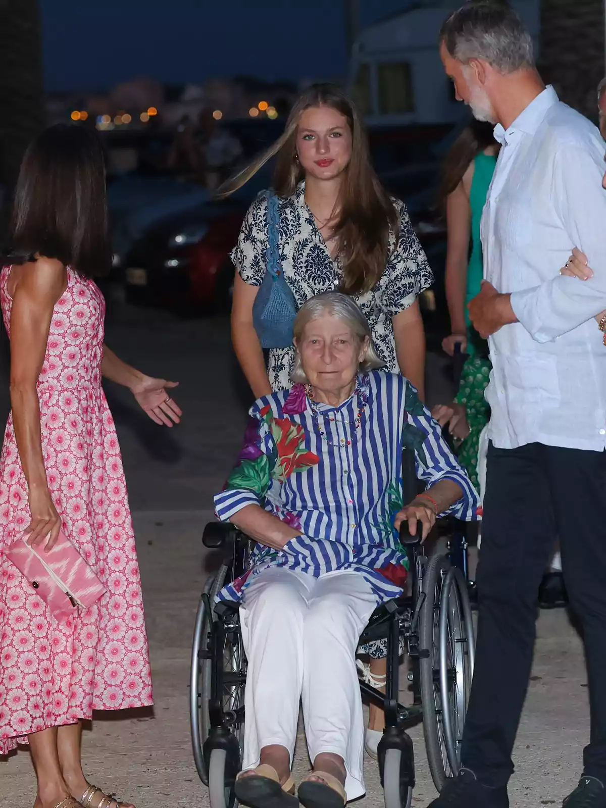 Una mujer mayor en silla de ruedas está rodeada de tres personas, dos mujeres y un hombre, en un ambiente al aire libre durante la noche Una mujer mayor en silla de ruedas está rodeada de tres personas, dos mujeres y un hombre, en un ambiente al aire libre durante la noche