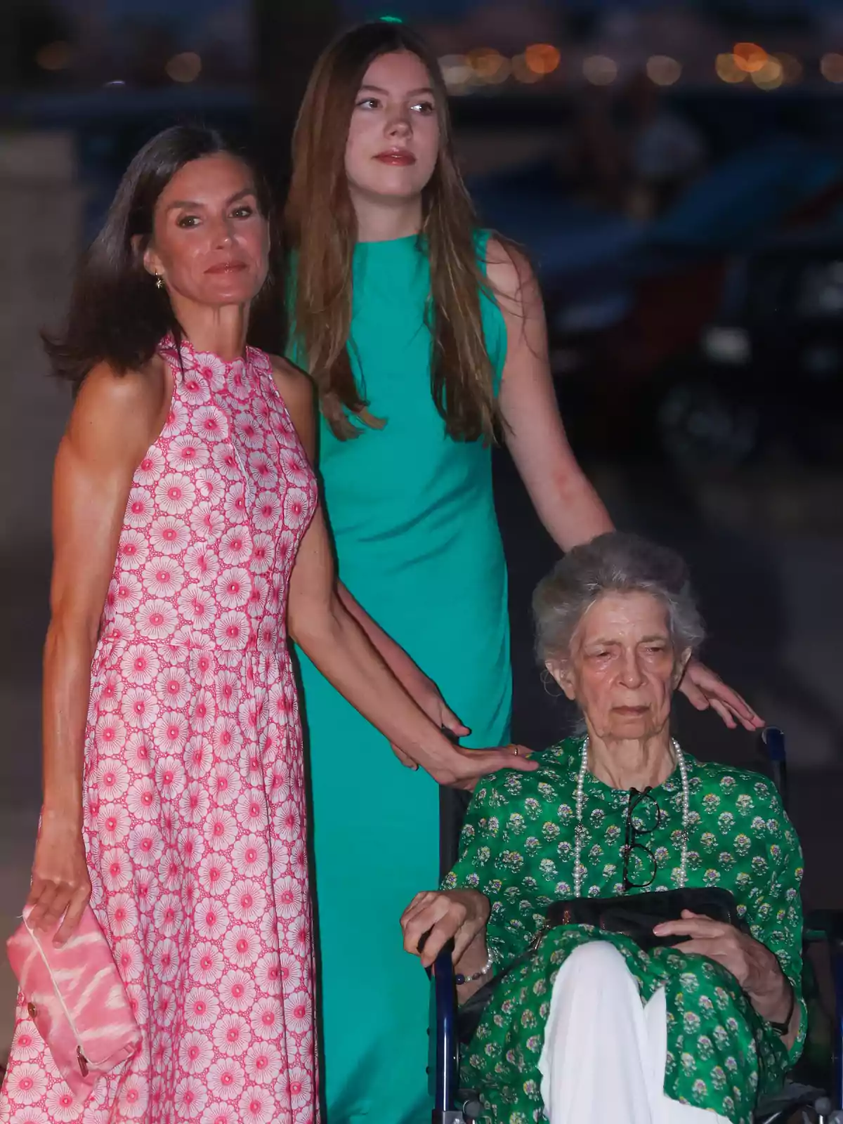 Tres mujeres de diferentes generaciones posan juntas al aire libre durante la noche, una de ellas está sentada en silla de ruedas y las otras dos de pie a su lado, todas lucen vestidos coloridos Tres mujeres de diferentes generaciones posan juntas al aire libre durante la noche, una de ellas está sentada en silla de ruedas y las otras dos de pie a su lado, todas lucen vestidos coloridos