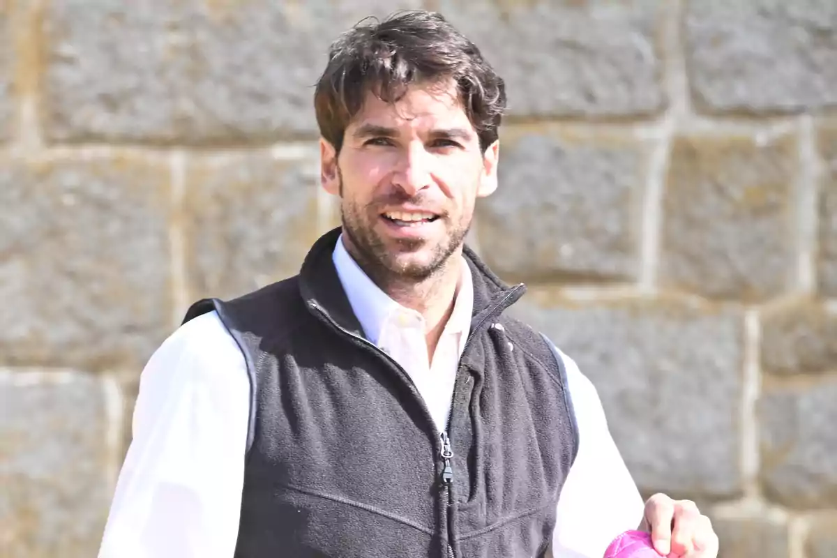 Hombre de cabello oscuro y barba corta vistiendo camisa blanca y chaleco negro frente a una pared de piedra