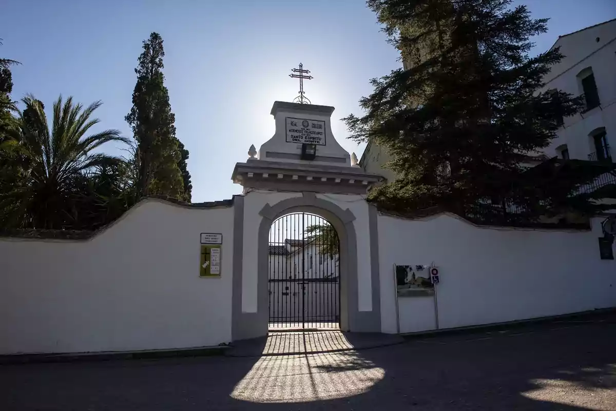 Entrada principal de un edificio religioso con una cruz en la parte superior, rodeada de árboles y con una reja proyectando sombra en el suelo.