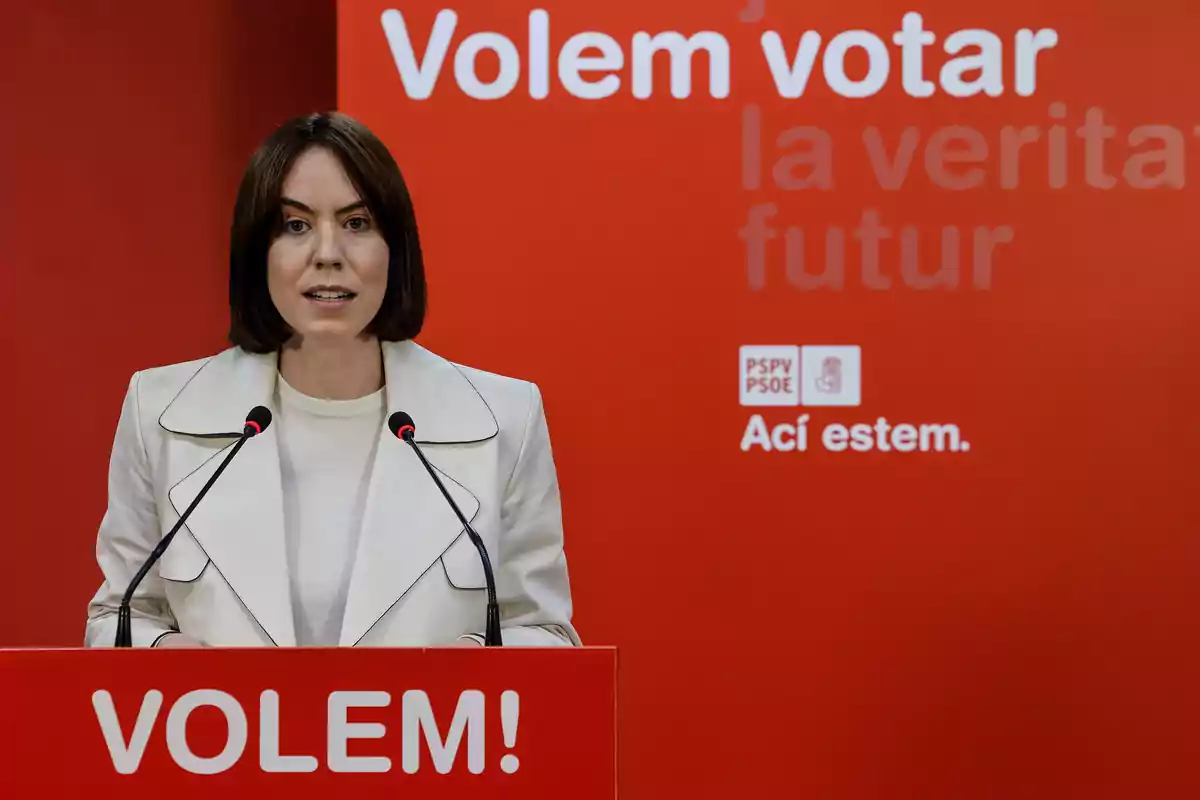 Mujer de cabello corto y oscuro hablando en un atril rojo con el texto VOLEM frente a un fondo rojo con mensajes en valenciano y logotipos del PSPV PSOE Mujer de cabello corto y oscuro hablando en un atril rojo con el texto VOLEM frente a un fondo rojo con mensajes en valenciano y logotipos del PSPV PSOE