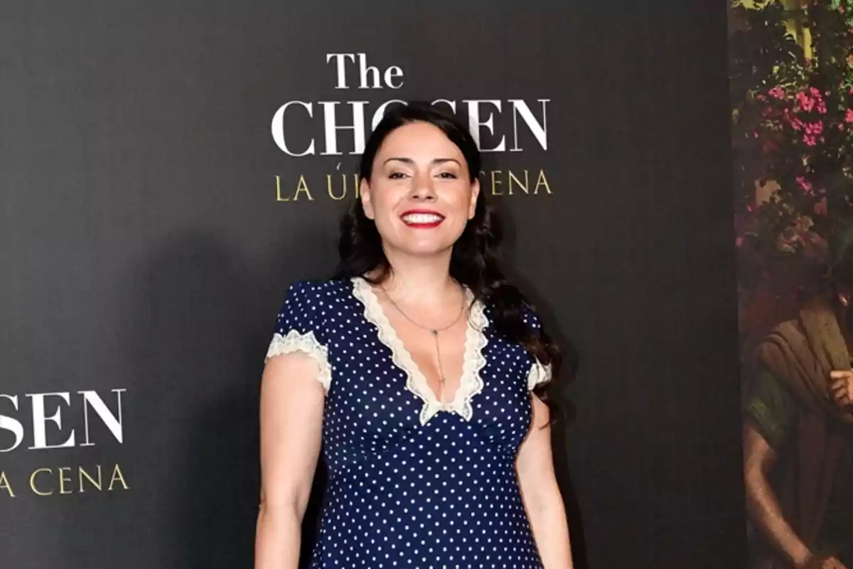 Mujer de cabello oscuro y vestido azul con lunares blancos posando sonriente frente a un fondo promocional de la serie The Chosen La Última Cena Mujer de cabello oscuro y vestido azul con lunares blancos posando sonriente frente a un fondo promocional de la serie The Chosen La Última Cena