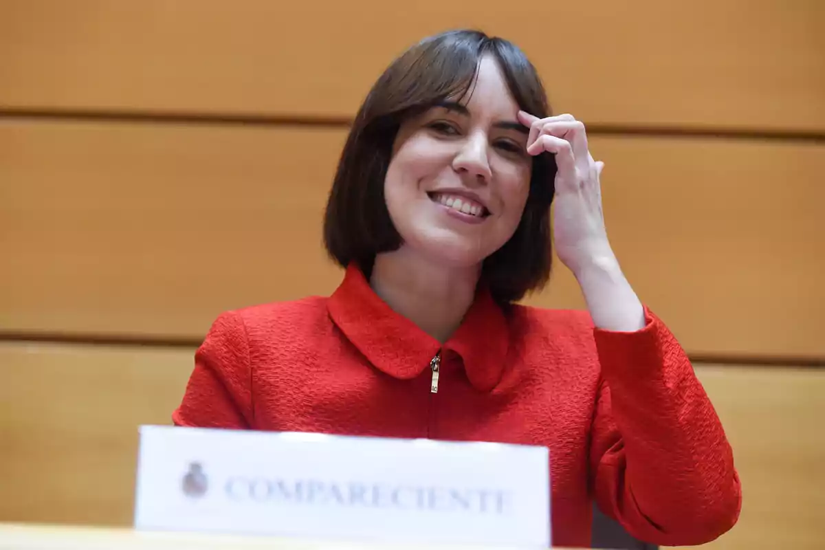 Mujer de cabello corto y oscuro con chaqueta roja sonriendo y sentada frente a un cartel que dice compareciente en un entorno institucional Mujer de cabello corto y oscuro con chaqueta roja sonriendo y sentada frente a un cartel que dice compareciente en un entorno institucional