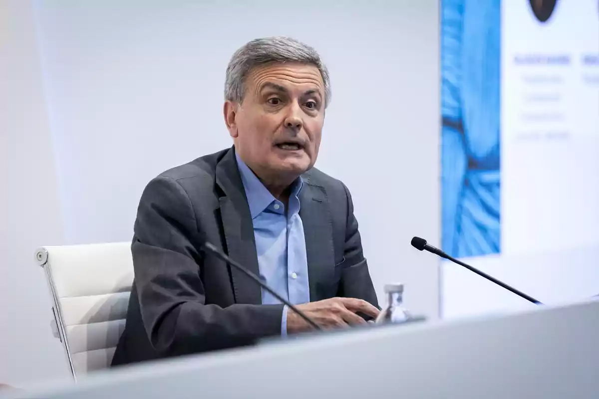 Hombre de cabello canoso con traje gris y camisa azul hablando en una conferencia sentado frente a un micrófono Hombre de cabello canoso con traje gris y camisa azul hablando en una conferencia sentado frente a un micrófono