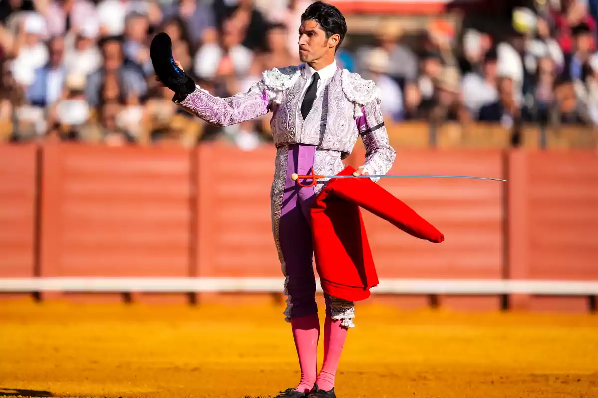 Torero vestido con traje tradicional sostiene una montera en una mano y una muleta roja en la otra en una plaza de toros frente al público.