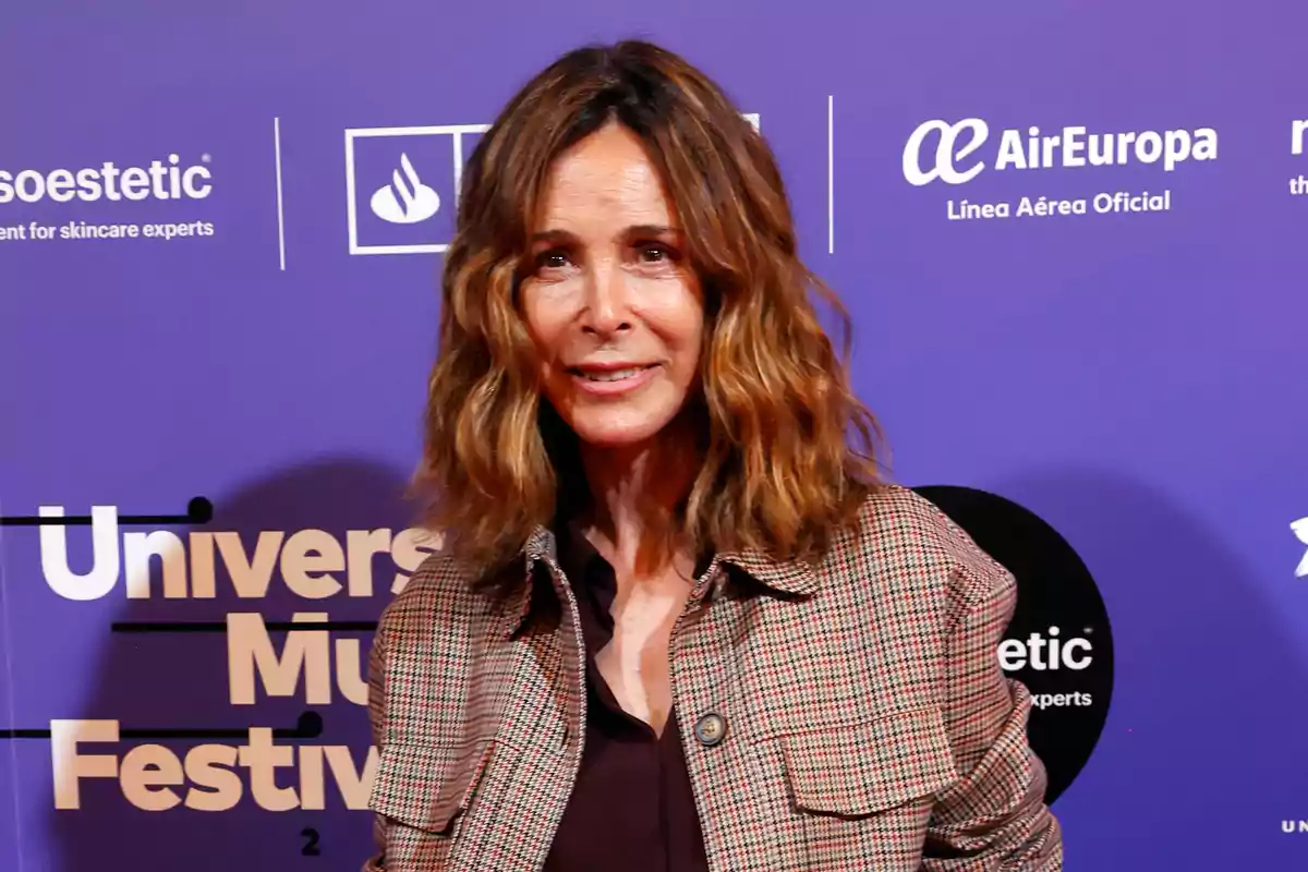 Mujer de cabello castaño ondulado y suelto posando frente a un fondo morado con logotipos y texto de un evento