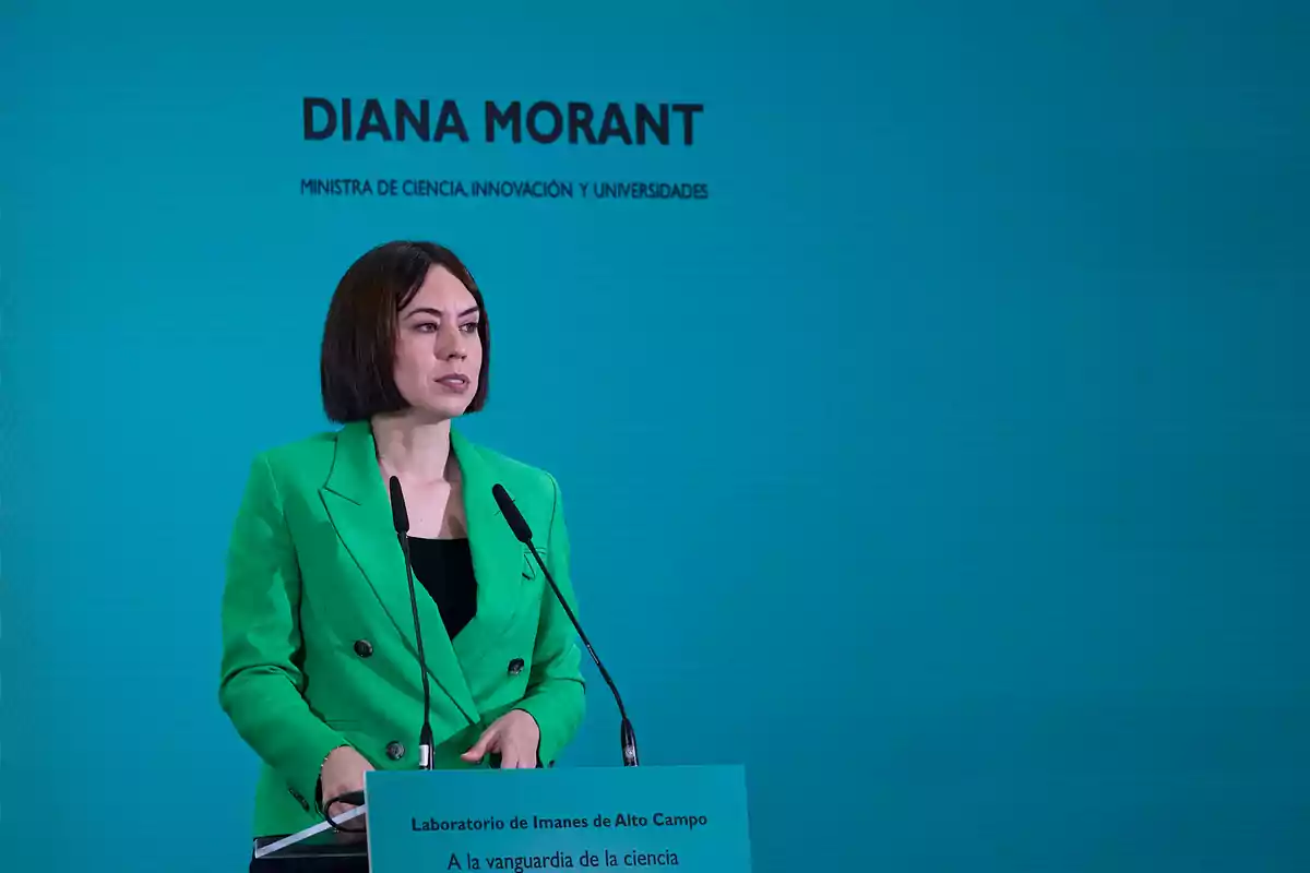 Mujer con chaqueta verde hablando en un atril frente a un fondo azul con texto institucional Mujer con chaqueta verde hablando en un atril frente a un fondo azul con texto institucional