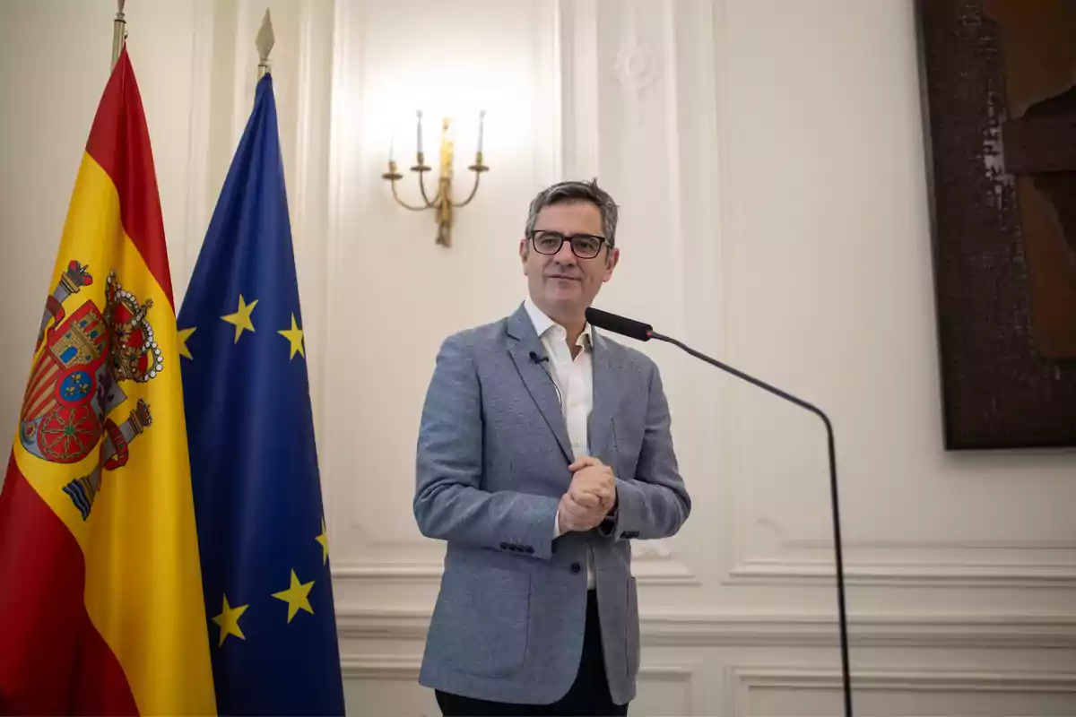 Hombre con gafas y chaqueta gris hablando frente a un micrófono junto a las banderas de España y la Unión Europea en una sala elegante Hombre con gafas y chaqueta gris hablando frente a un micrófono junto a las banderas de España y la Unión Europea en una sala elegante