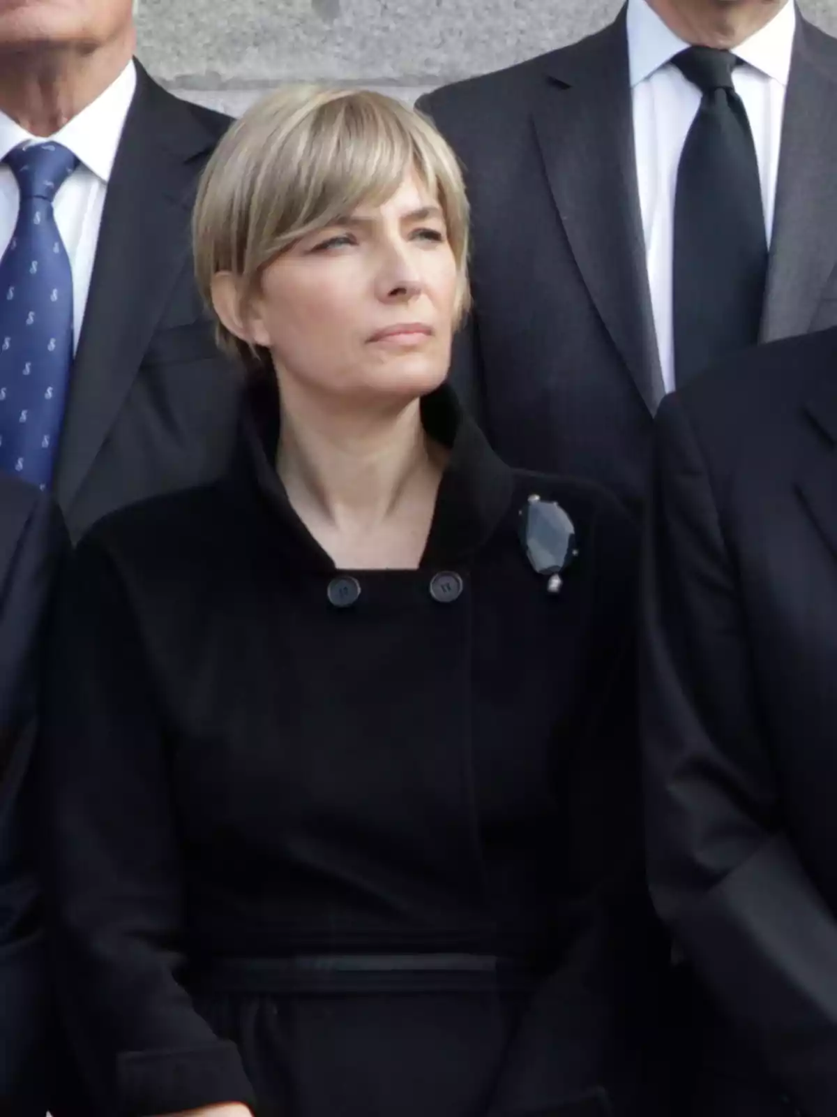 Mujer de cabello corto rubio vestida de negro, de pie entre personas con trajes formales