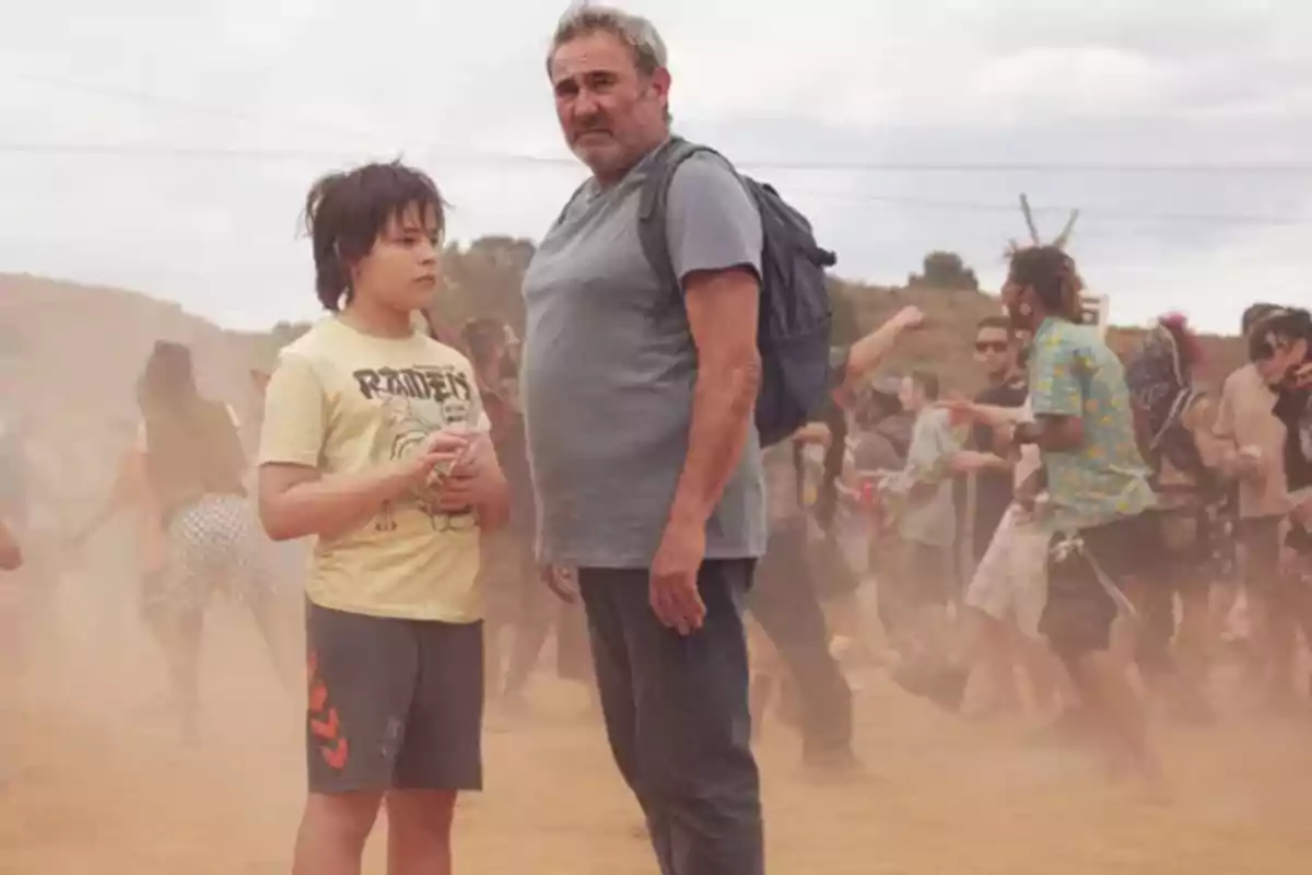 Un hombre mayor con mochila y un niño están de pie en medio de un grupo de personas en un ambiente polvoriento al aire libre Un hombre mayor con mochila y un niño están de pie en medio de un grupo de personas en un ambiente polvoriento al aire libre
