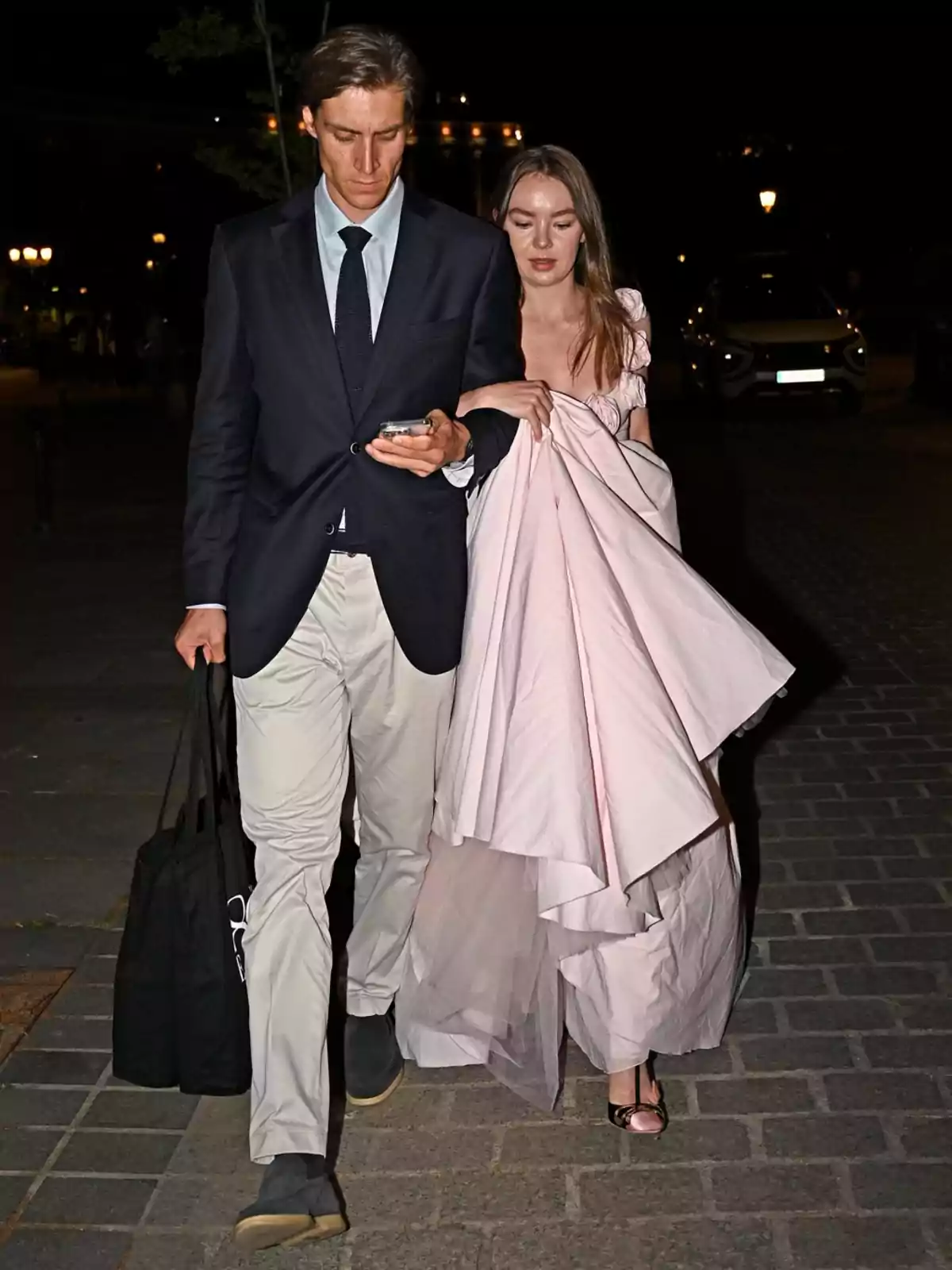 Pareja elegante caminando de noche por la calle, él lleva traje oscuro y ella un vestido largo rosa claro