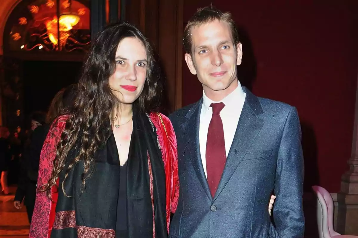 Una pareja posa sonriente en un evento elegante, ella lleva un abrigo rojo y bufanda negra, él viste traje azul con corbata roja