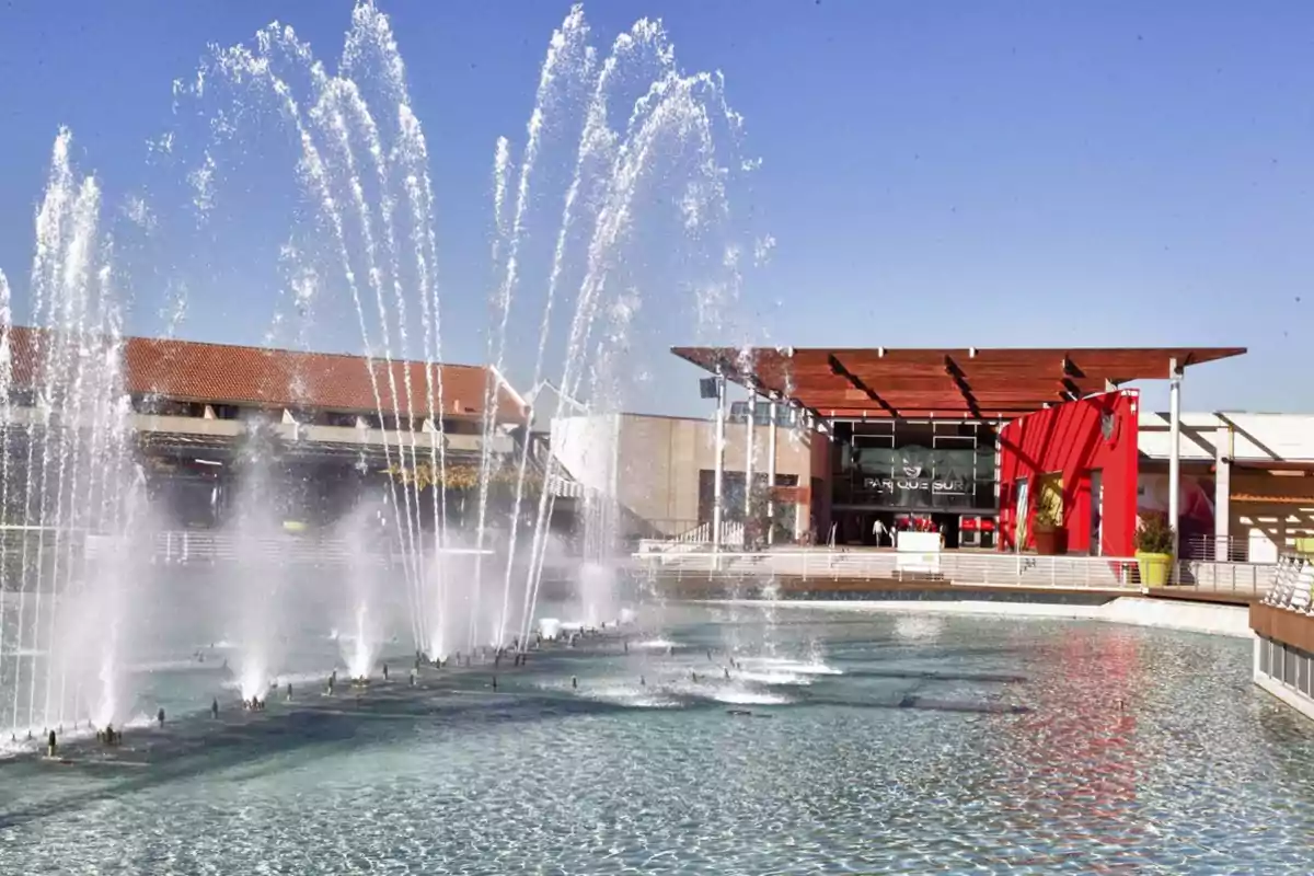 Fuente de agua con chorros altos frente a un centro comercial moderno bajo un cielo despejado Fuente de agua con chorros altos frente a un centro comercial moderno bajo un cielo despejado