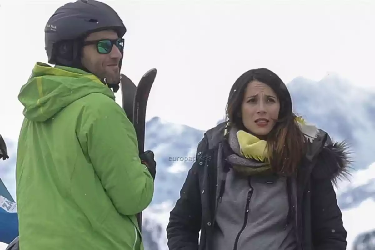 Dos personas con ropa de invierno en una estación de esquí, una de ellas lleva casco y gafas de sol mientras sostiene unos esquís, la otra viste bufanda y chaqueta con capucha de peluche, al fondo se ven montañas nevadas Dos personas con ropa de invierno en una estación de esquí, una de ellas lleva casco y gafas de sol mientras sostiene unos esquís, la otra viste bufanda y chaqueta con capucha de peluche, al fondo se ven montañas nevadas