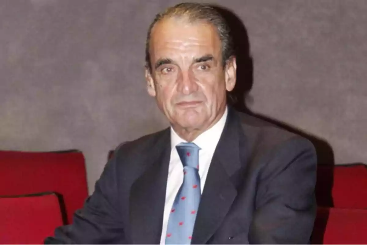 Hombre de cabello canoso peinado hacia atrás vistiendo traje oscuro y corbata azul sentado en una silla roja Hombre de cabello canoso peinado hacia atrás vistiendo traje oscuro y corbata azul sentado en una silla roja