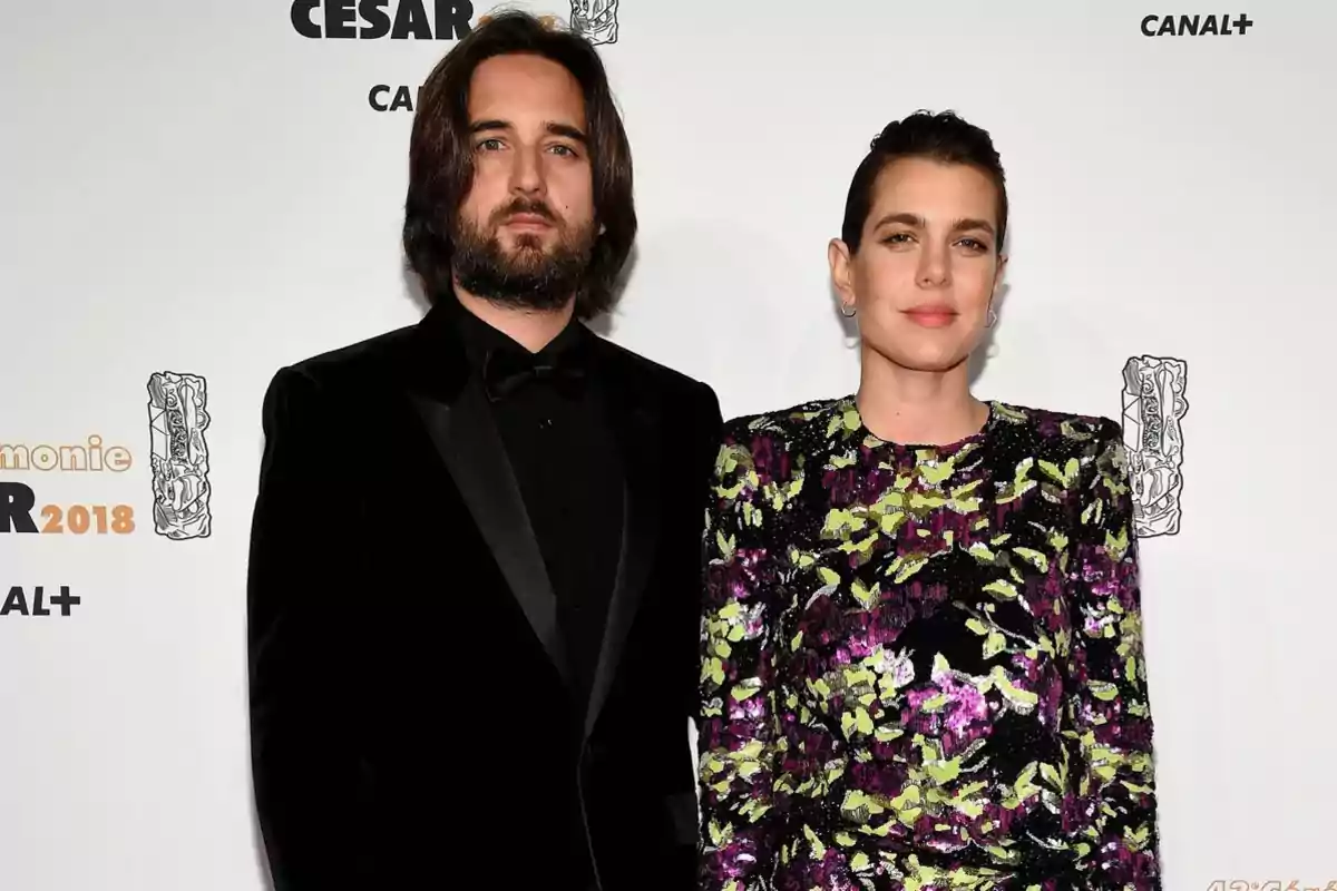 Una pareja posa en una alfombra roja frente a un fondo con logotipos de un evento de 2018, él lleva esmoquin negro y ella un vestido de colores con estampado floral.
