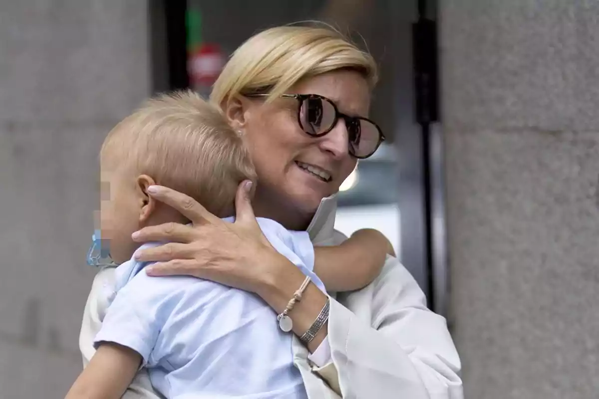 Mujer rubia con gafas de sol abrazando a un niño pequeño rubio con chupete frente a una pared gris Mujer rubia con gafas de sol abrazando a un niño pequeño rubio con chupete frente a una pared gris