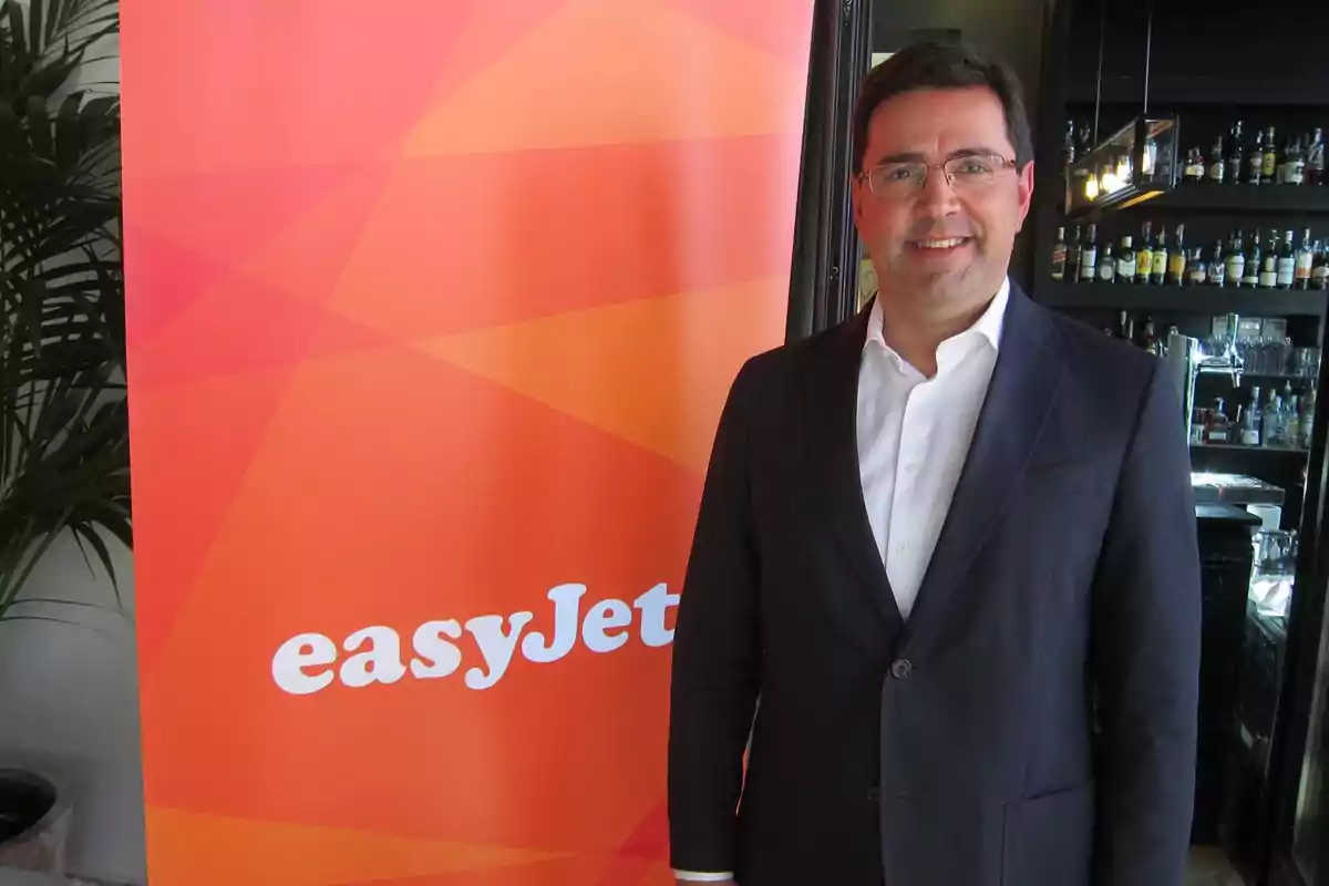 Hombre con gafas y traje oscuro sonríe junto a un cartel naranja de easyJet en un entorno interior con botellas al fondo. Hombre con gafas y traje oscuro sonríe junto a un cartel naranja de easyJet en un entorno interior con botellas al fondo.