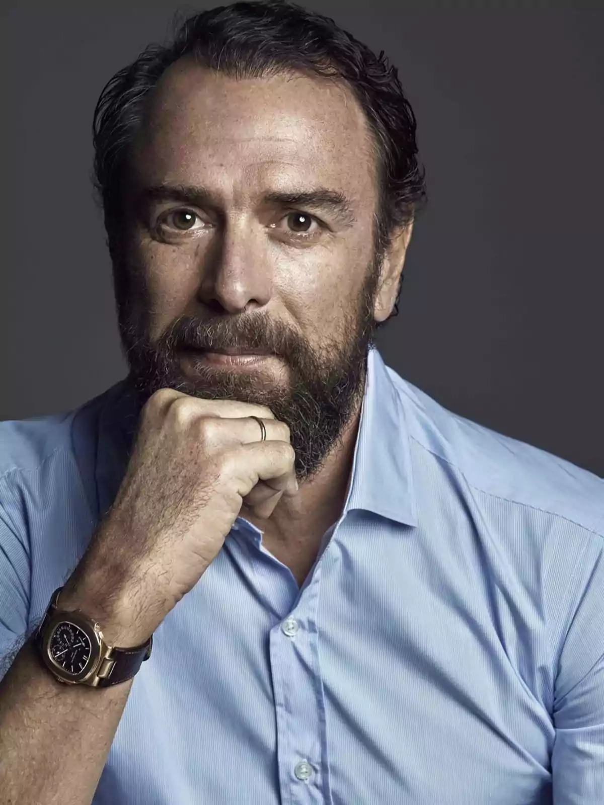 Hombre de barba y cabello oscuro con camisa azul claro y reloj dorado apoyando el mentón sobre su mano Hombre de barba y cabello oscuro con camisa azul claro y reloj dorado apoyando el mentón sobre su mano