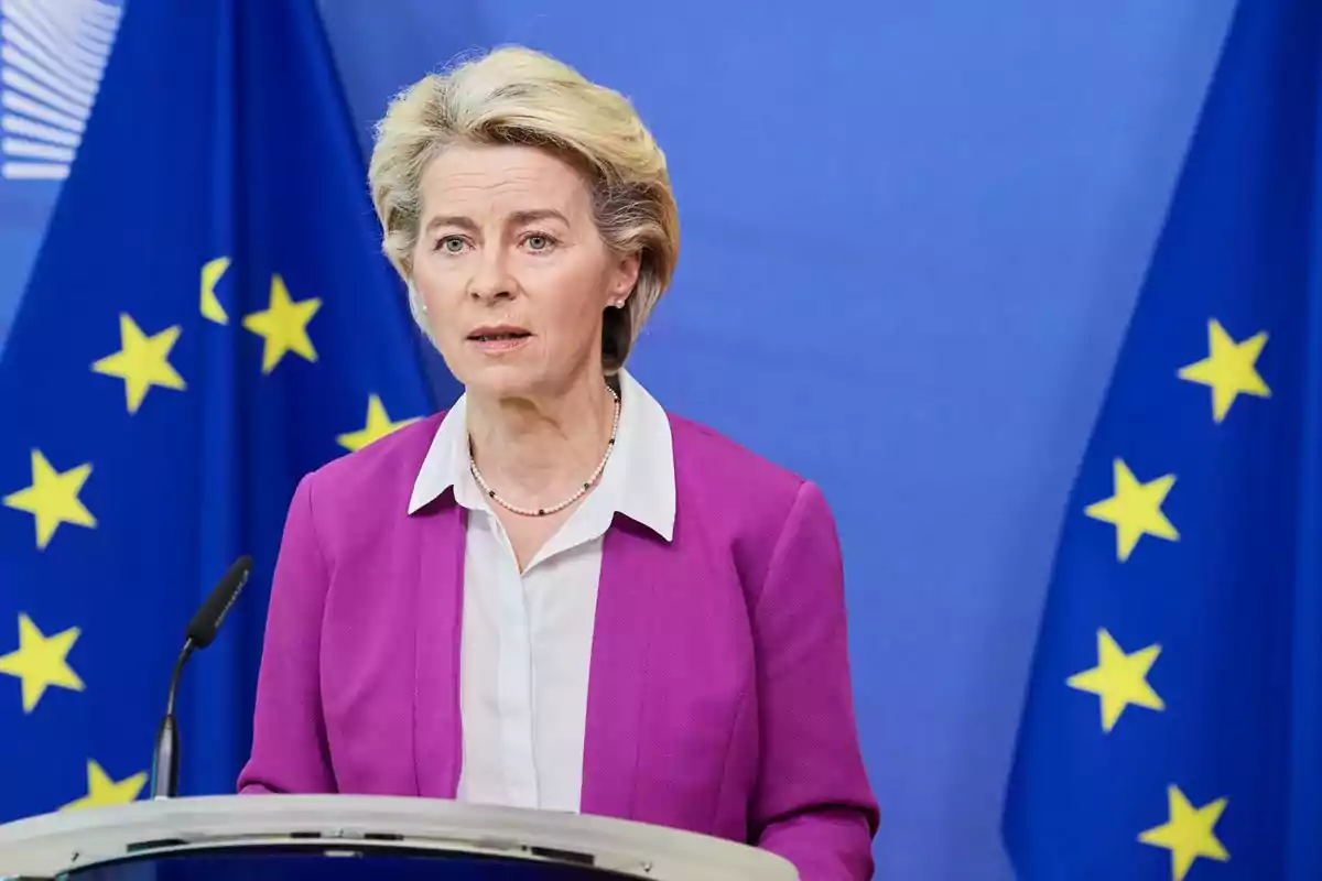 Mujer de cabello rubio con saco fucsia hablando frente a un micrófono con banderas de la Unión Europea de fondo Mujer de cabello rubio con saco fucsia hablando frente a un micrófono con banderas de la Unión Europea de fondo