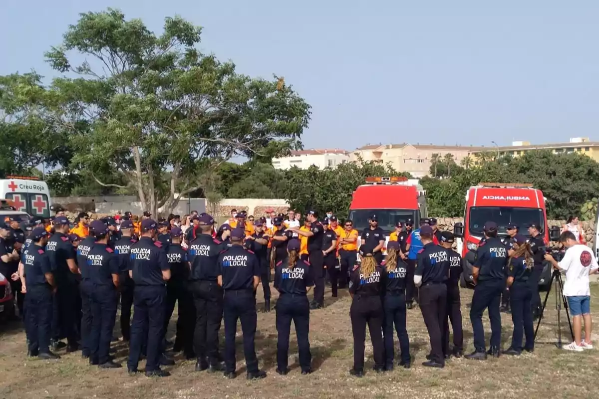 Un grupo numeroso de policías locales y personal de emergencias se reúne al aire libre frente a ambulancias y vehículos de la Cruz Roja en una zona urbana con árboles y edificios al fondo
