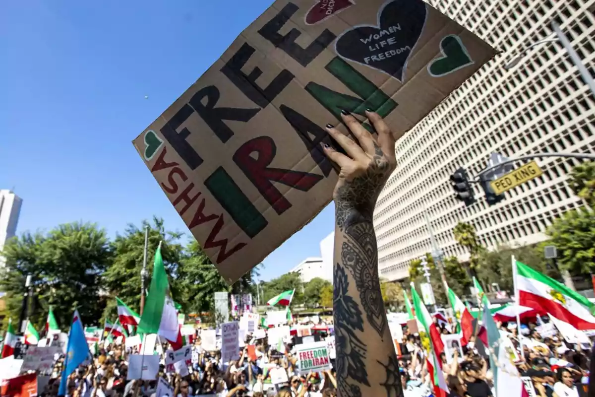 Persona con tatuajes en el brazo sostiene un cartel que dice Free Iran durante una manifestación con muchas personas y banderas en la calle frente a un edificio grande Persona con tatuajes en el brazo sostiene un cartel que dice Free Iran durante una manifestación con muchas personas y banderas en la calle frente a un edificio grande
