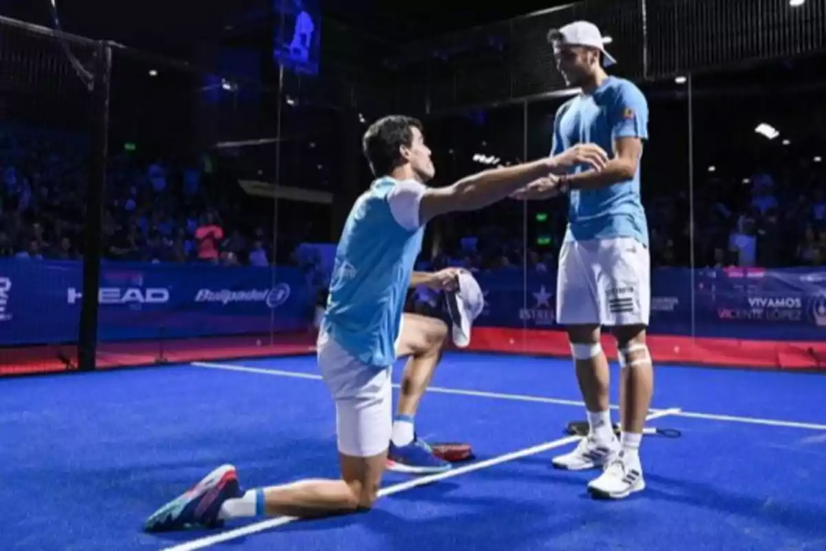 Dos jugadores de pádel en una cancha azul celebran un punto, uno de ellos está arrodillado extendiendo los brazos hacia su compañero que está de pie sonriendo Dos jugadores de pádel en una cancha azul celebran un punto, uno de ellos está arrodillado extendiendo los brazos hacia su compañero que está de pie sonriendo