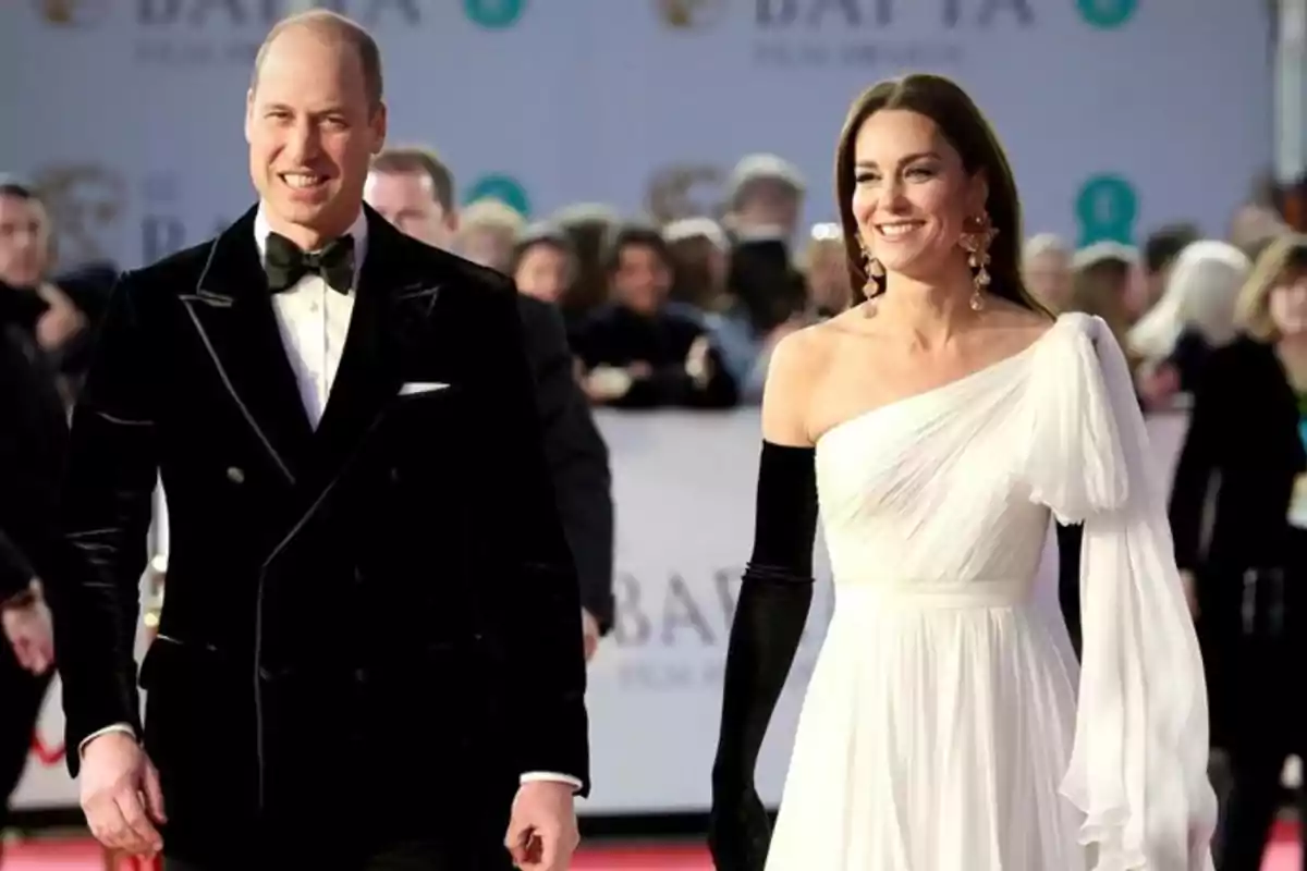 Pareja elegantemente vestida caminando por una alfombra roja rodeada de personas al fondo