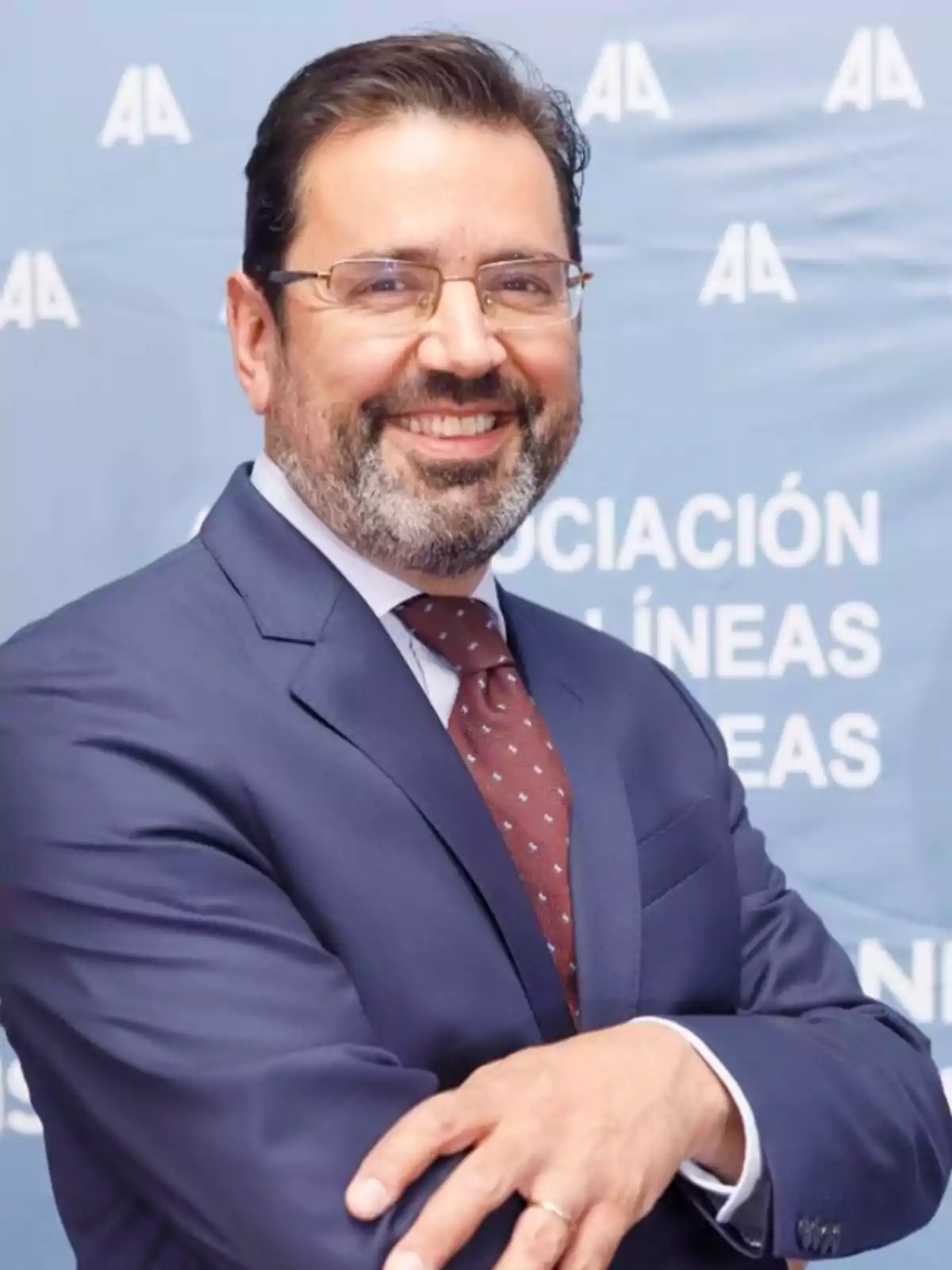 Hombre de mediana edad con barba y gafas, vestido con traje azul y corbata, sonriendo frente a un fondo azul con letras blancas Hombre de mediana edad con barba y gafas, vestido con traje azul y corbata, sonriendo frente a un fondo azul con letras blancas