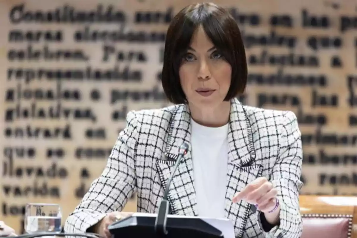 Mujer de cabello corto y oscuro con saco blanco de cuadros hablando frente a un micrófono en un entorno formal con fondo de texto borroso