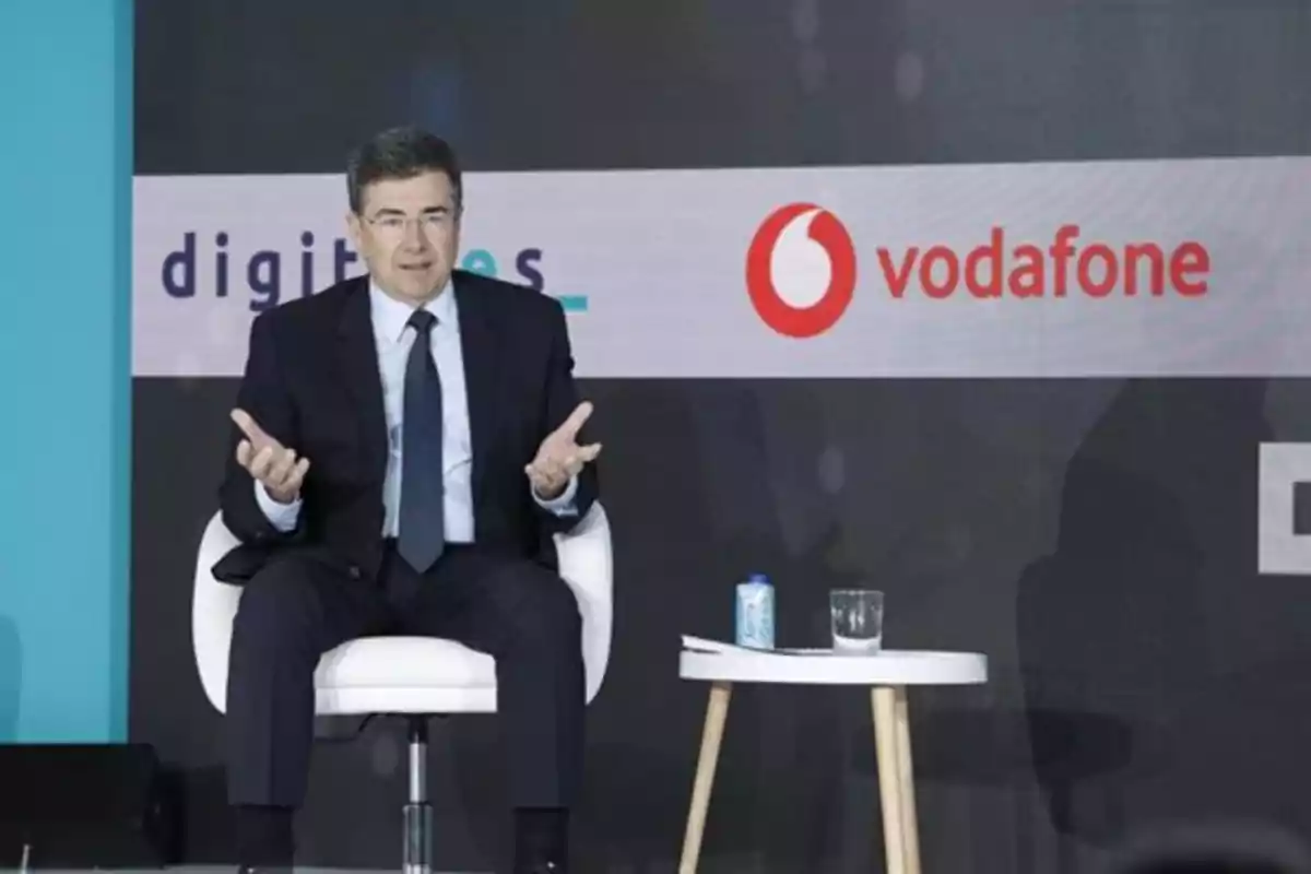 Hombre con traje sentado en una silla blanca participando en un evento con fondo de logotipos de empresas y una mesa pequeña con una lata y un vaso de agua a su lado Hombre con traje sentado en una silla blanca participando en un evento con fondo de logotipos de empresas y una mesa pequeña con una lata y un vaso de agua a su lado