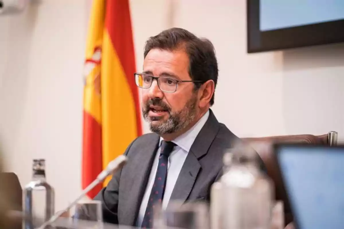 Hombre con gafas y barba hablando en una conferencia con una bandera de España al fondo Hombre con gafas y barba hablando en una conferencia con una bandera de España al fondo