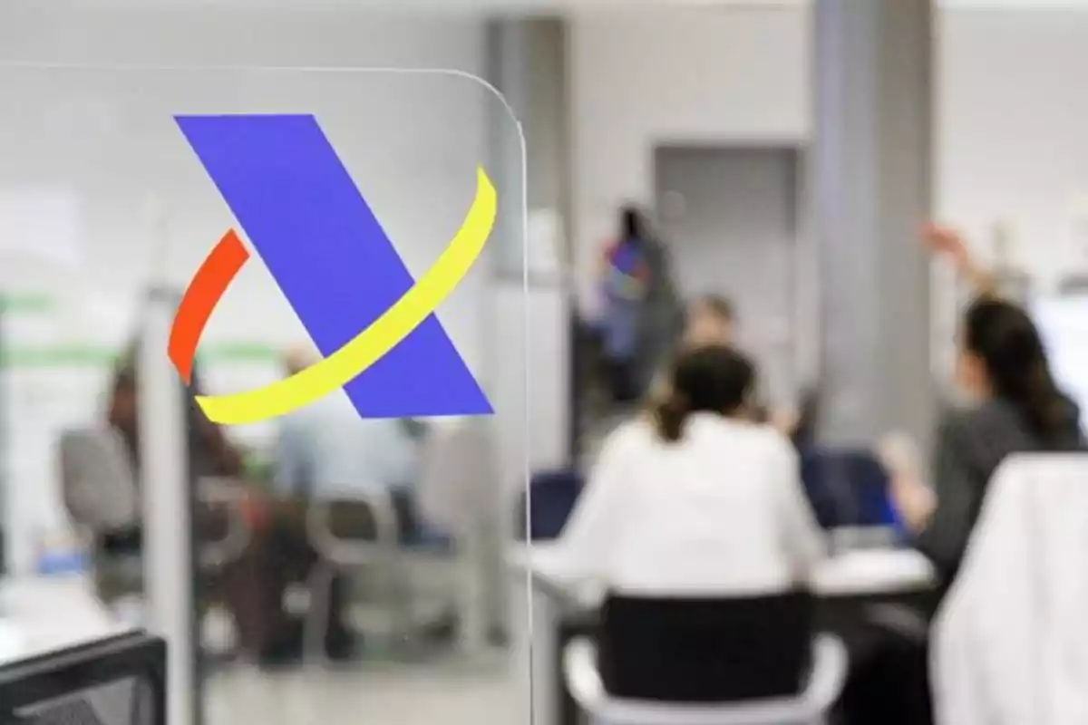 Logo de la Agencia Tributaria en una oficina con varias personas atendiendo trámites administrativos Logo de la Agencia Tributaria en una oficina con varias personas atendiendo trámites administrativos
