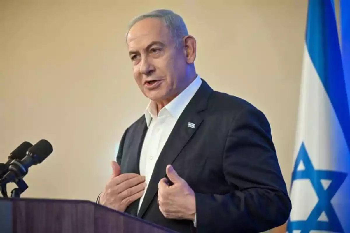 Hombre de cabello canoso con traje oscuro hablando frente a un micrófono y una bandera de Israel al fondo Hombre de cabello canoso con traje oscuro hablando frente a un micrófono y una bandera de Israel al fondo