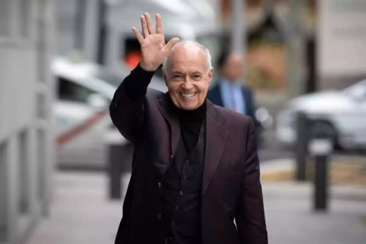 Hombre mayor sonriente con cabello canoso saluda levantando la mano mientras camina por la calle vestido con saco oscuro Hombre mayor sonriente con cabello canoso saluda levantando la mano mientras camina por la calle vestido con saco oscuro