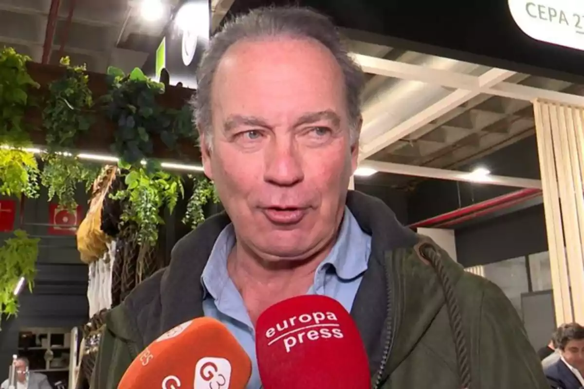Un hombre de mediana edad con cabello canoso está hablando frente a varios micrófonos de prensa en un evento interior con plantas decorativas al fondo. Un hombre de mediana edad con cabello canoso está hablando frente a varios micrófonos de prensa en un evento interior con plantas decorativas al fondo.