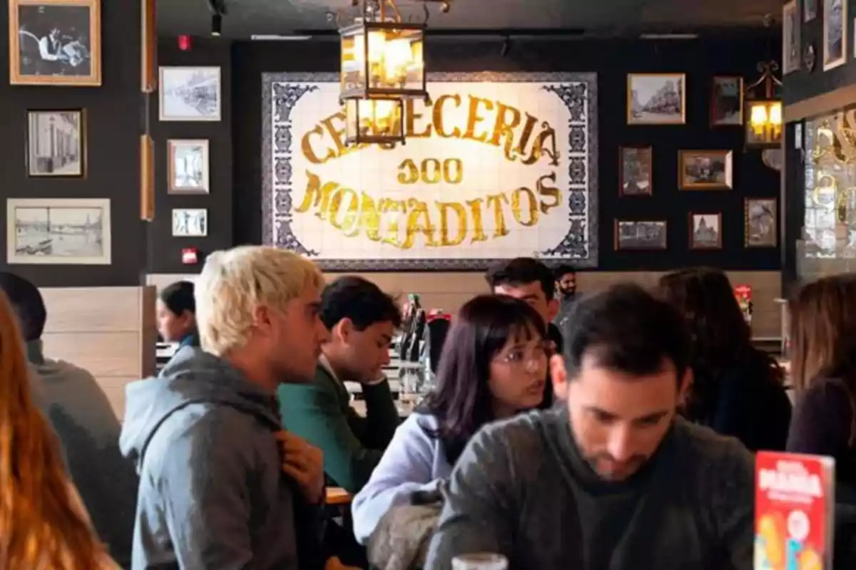 Personas sentadas conversando y comiendo en el interior de un restaurante decorado con cuadros y un gran letrero que dice cervecería 100 montaditos Personas sentadas conversando y comiendo en el interior de un restaurante decorado con cuadros y un gran letrero que dice cervecería 100 montaditos
