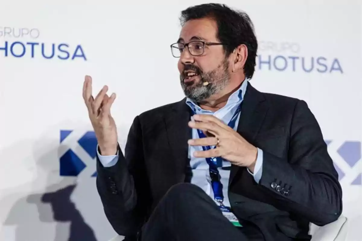 Hombre con barba y gafas hablando animadamente durante un evento empresarial sentado frente a un fondo con el logo de Grupo Hotusa Hombre con barba y gafas hablando animadamente durante un evento empresarial sentado frente a un fondo con el logo de Grupo Hotusa