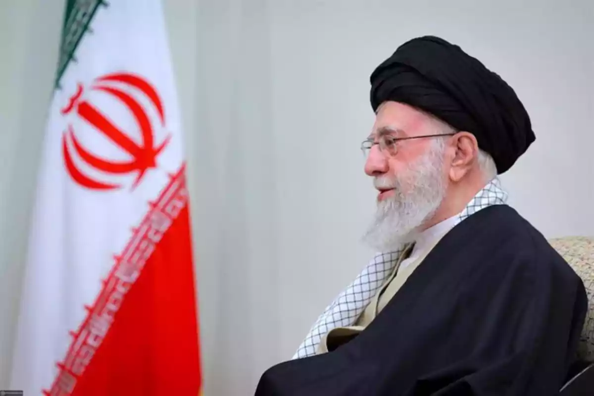 Hombre mayor con barba y turbante negro sentado junto a una bandera de Irán Hombre mayor con barba y turbante negro sentado junto a una bandera de Irán