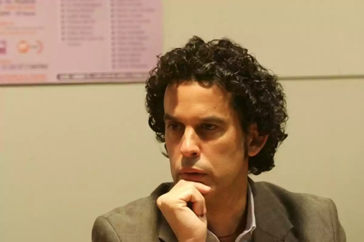 Hombre de cabello rizado y oscuro con expresión pensativa apoyando la barbilla en su mano, viste saco y camisa clara, fondo desenfocado. Hombre de cabello rizado y oscuro con expresión pensativa apoyando la barbilla en su mano, viste saco y camisa clara, fondo desenfocado.