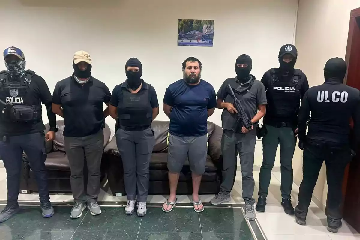 Un grupo de policías con uniforme y pasamontañas escoltan a un hombre con barba y ropa casual en una sala con sillones. Un grupo de policías con uniforme y pasamontañas escoltan a un hombre con barba y ropa casual en una sala con sillones.