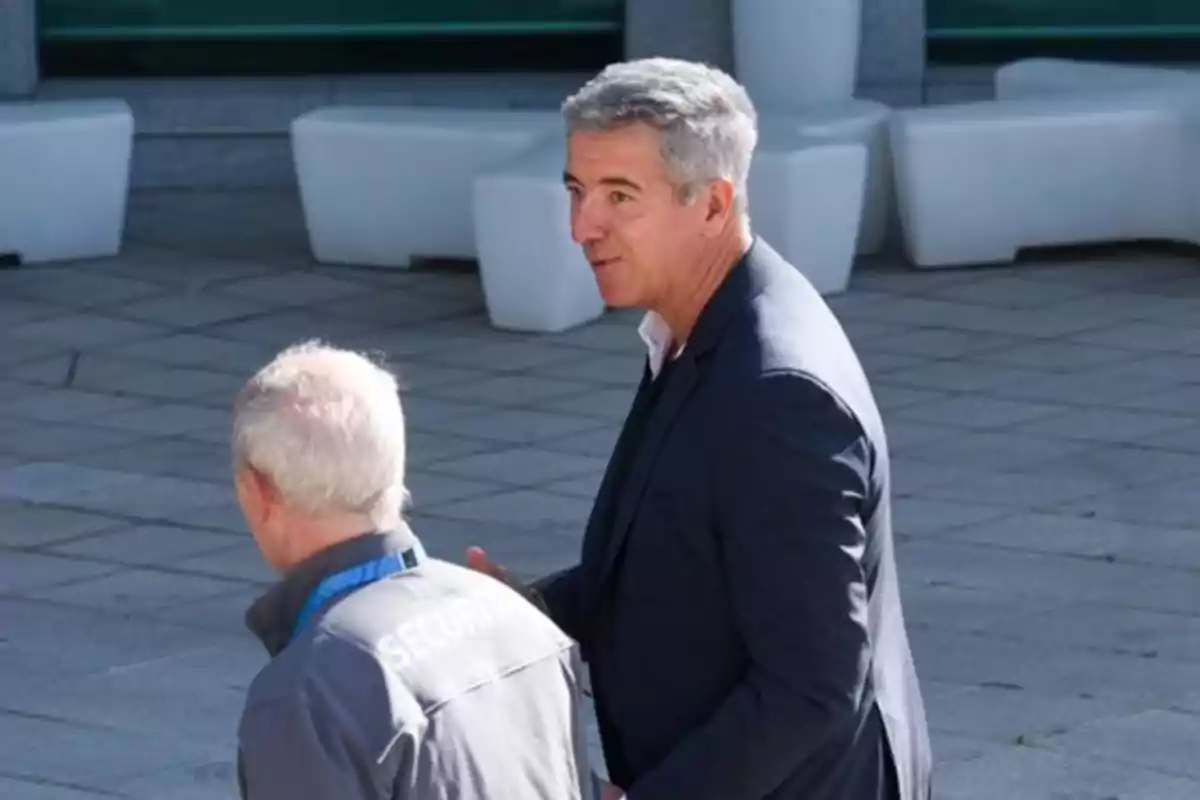 Dos hombres conversan al aire libre uno de ellos lleva un uniforme de seguridad y el otro viste un saco oscuro Dos hombres conversan al aire libre uno de ellos lleva un uniforme de seguridad y el otro viste un saco oscuro