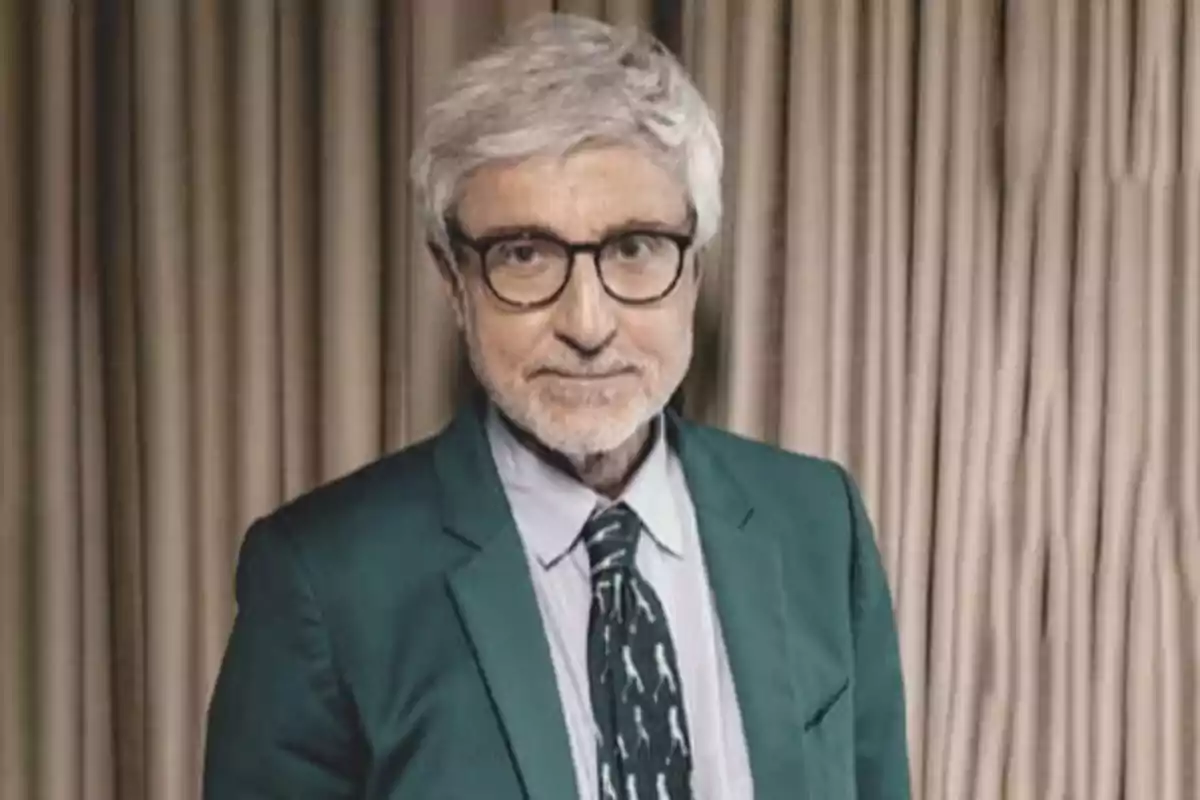 Hombre de cabello canoso y gafas con saco verde y corbata, posando frente a una cortina beige Hombre de cabello canoso y gafas con saco verde y corbata, posando frente a una cortina beige