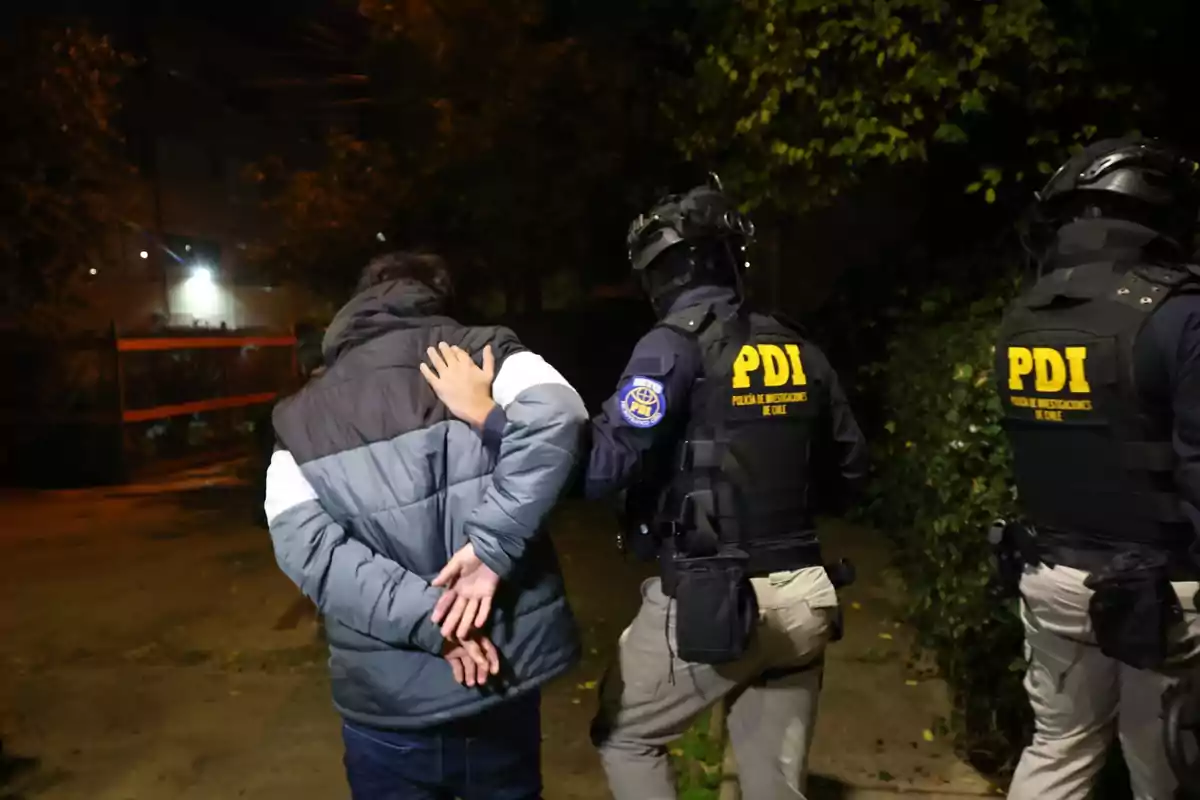 Un hombre es escoltado por dos agentes de la PDI durante la noche en una calle rodeada de vegetación.