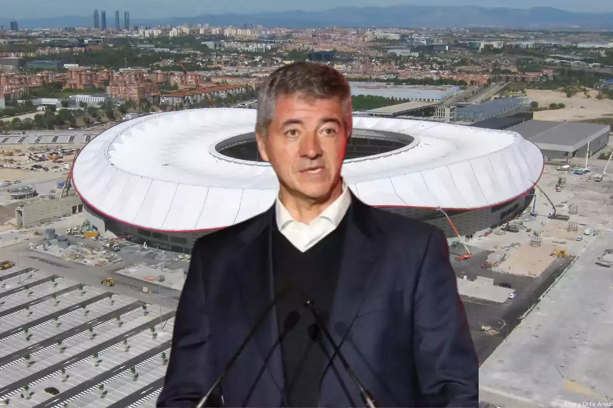 Un hombre de cabello canoso con traje oscuro está hablando frente a un estadio moderno de gran tamaño en una ciudad.