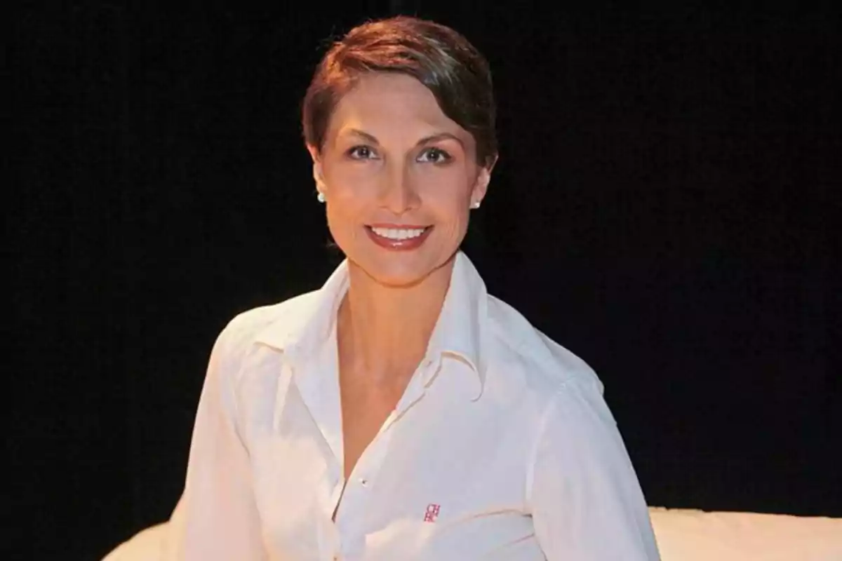Mujer de cabello corto y castaño sonriendo, vestida con camisa blanca y fondo oscuro