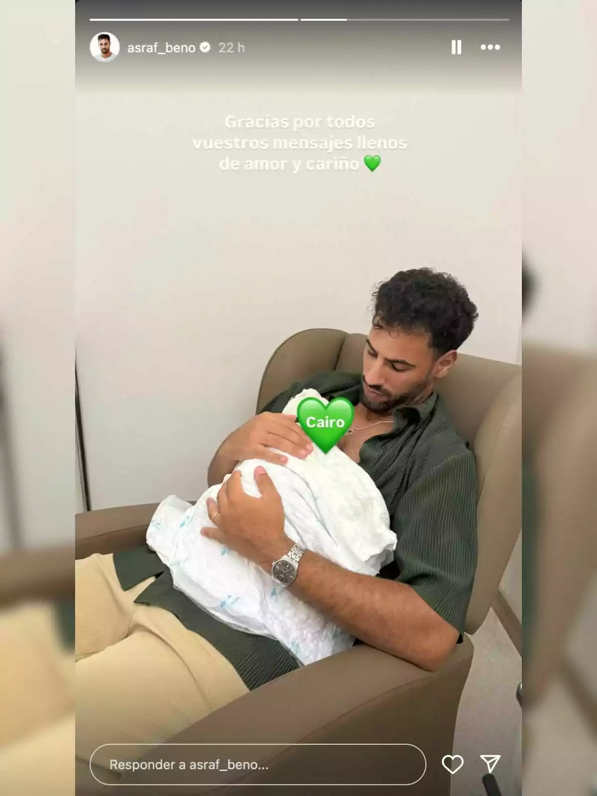 Asraf Beno sentado en un sillón sosteniendo a un bebé envuelto en una manta blanca con un emoji de corazón verde que cubre el rostro del bebé y la palabra Cairo escrita en el emoji. Asraf Beno sentado en un sillón sosteniendo a un bebé envuelto en una manta blanca con un emoji de corazón verde que cubre el rostro del bebé y la palabra Cairo escrita en el emoji.